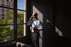 Hombre de negocios hablando por teléfono junto a una ventana, sosteniendo una taza.